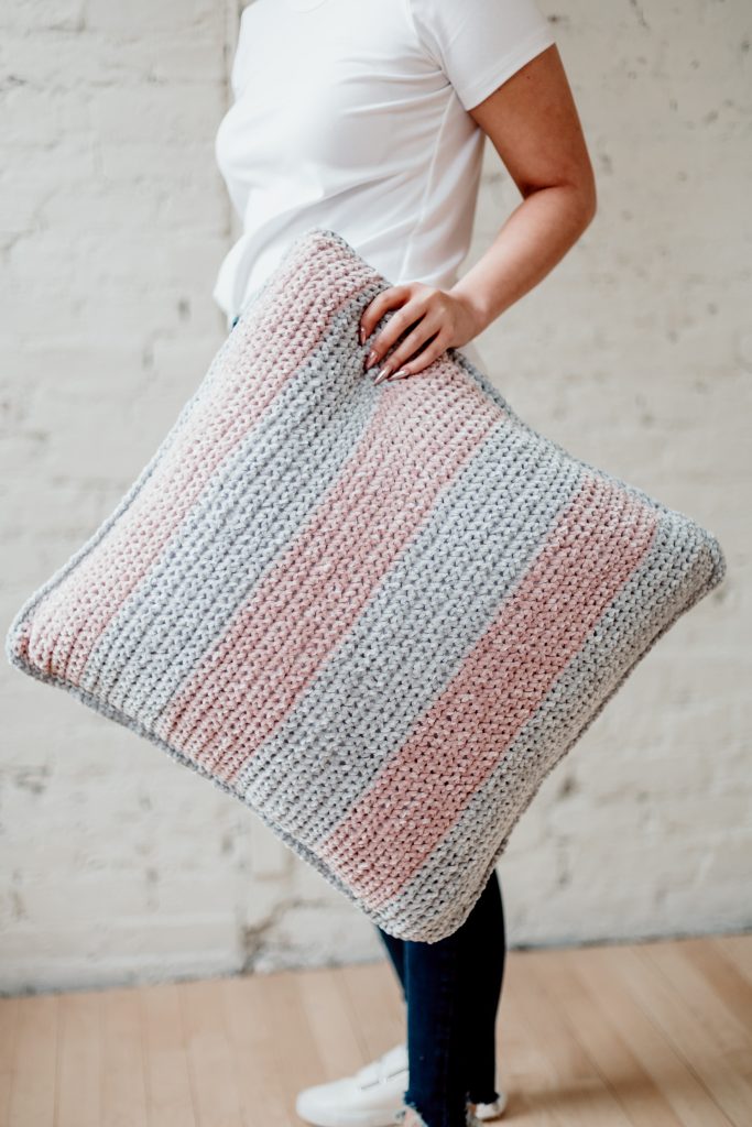 Spruce up your boring home decor with the Maisy Velvet pillow, a FREE beginner-friendly oversized throw pillow pattern from TLYCBlog. Pick your favorite velvet (chenille) yarn in three coordinating colors and bring together broad stripes and subtle details to give your living room a much-needed refresh. Perfect for beginners, this pattern uses simple stitches and works up quickly. Get the FREE pattern on the blog along with details on yarn choice and size. | TLYCBlog.com. 