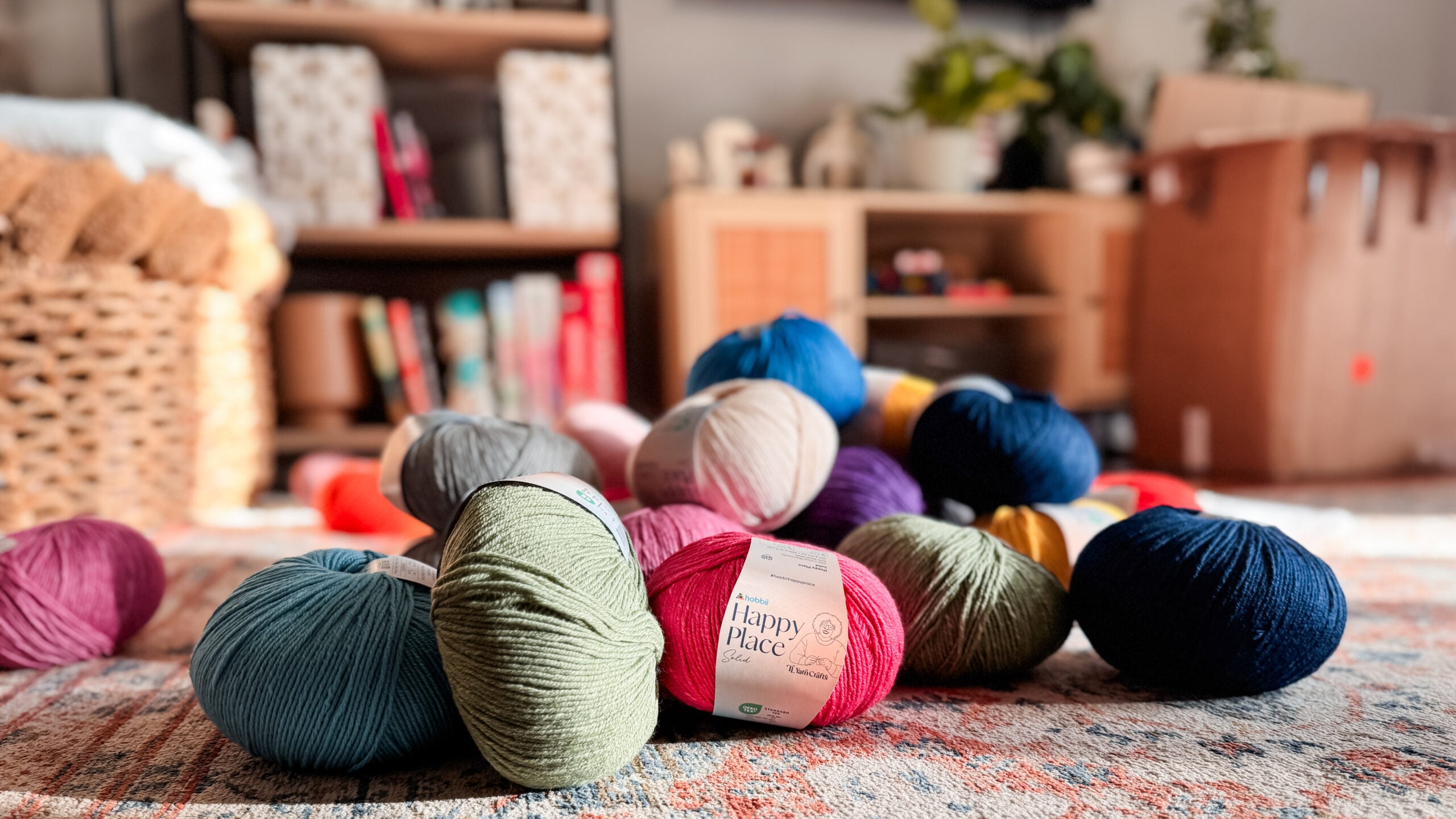 Assorted Happy Place yarn skeins in rich, saturated colors arranged on a rug with shelves and baskets in the background.