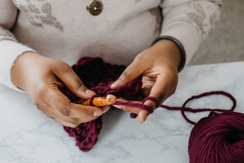 Toni Lipsey crocheting with bulky burgundy yarn and a large wooden crochet hook during a cozy crochet session.