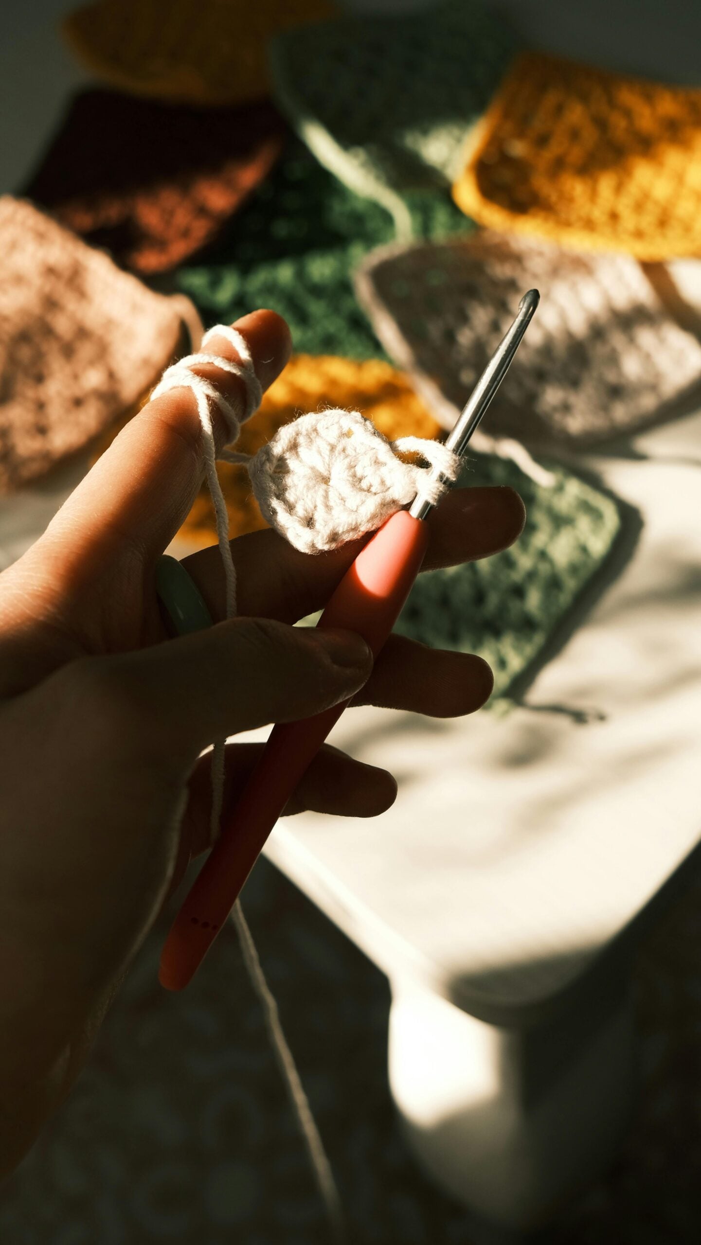 Hand holding a crochet hook while working a small granny square, with finished crochet squares in soft colors in the background.