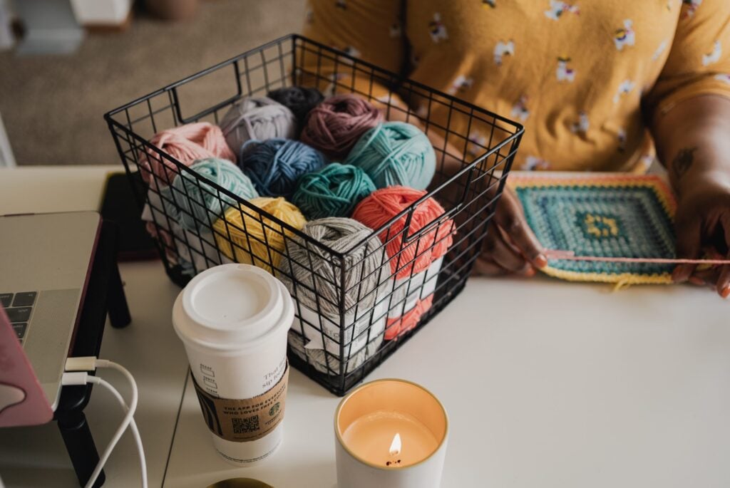 Crocheter working on a colorful granny square beside a basket of yarn, laptop, coffee, and candle.