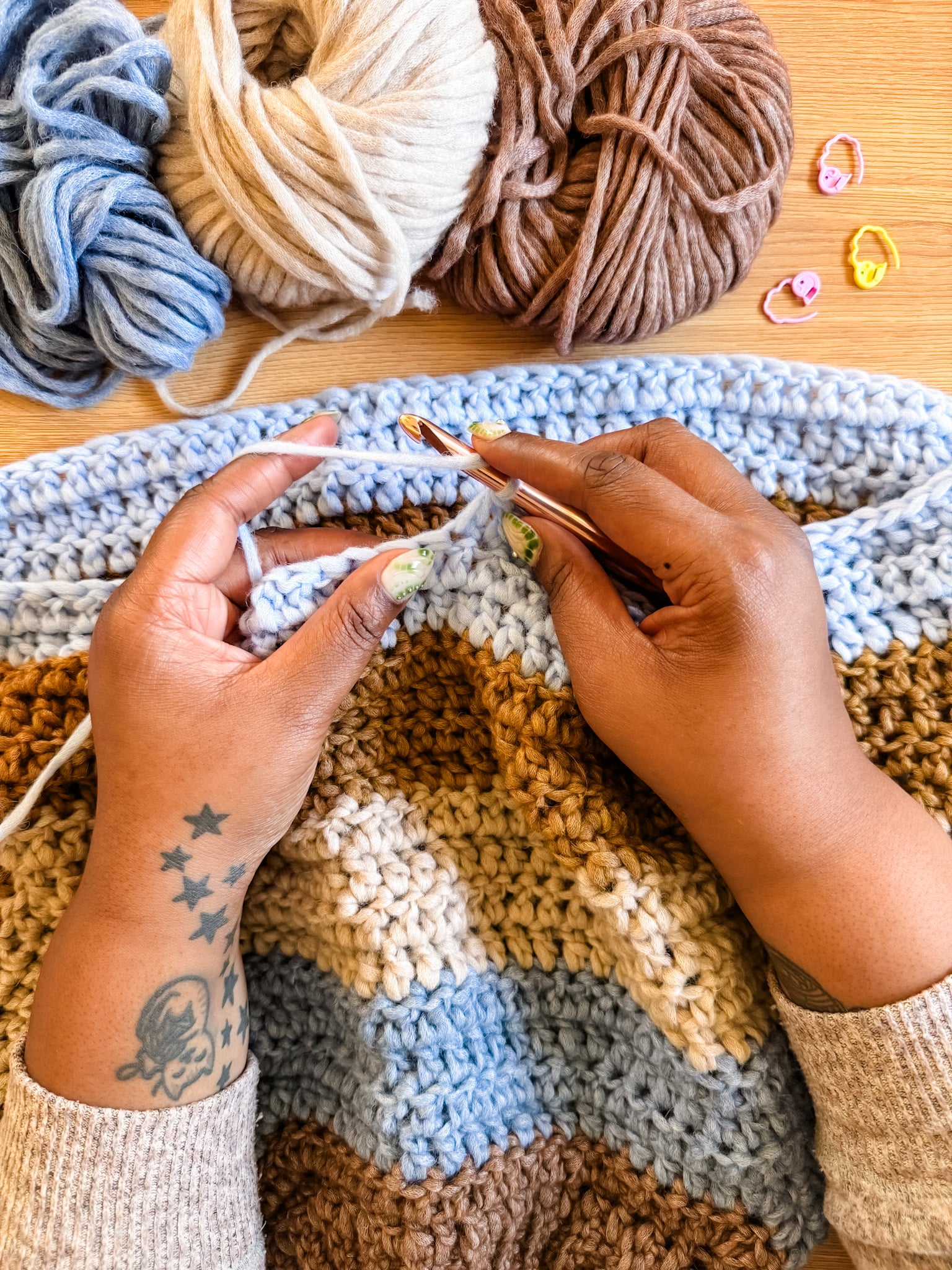 3 – Cover Image Hands crocheting a chunky crochet sweater in blue, brown, cream and gray, with coordinating yarn and stitch markers nearby.