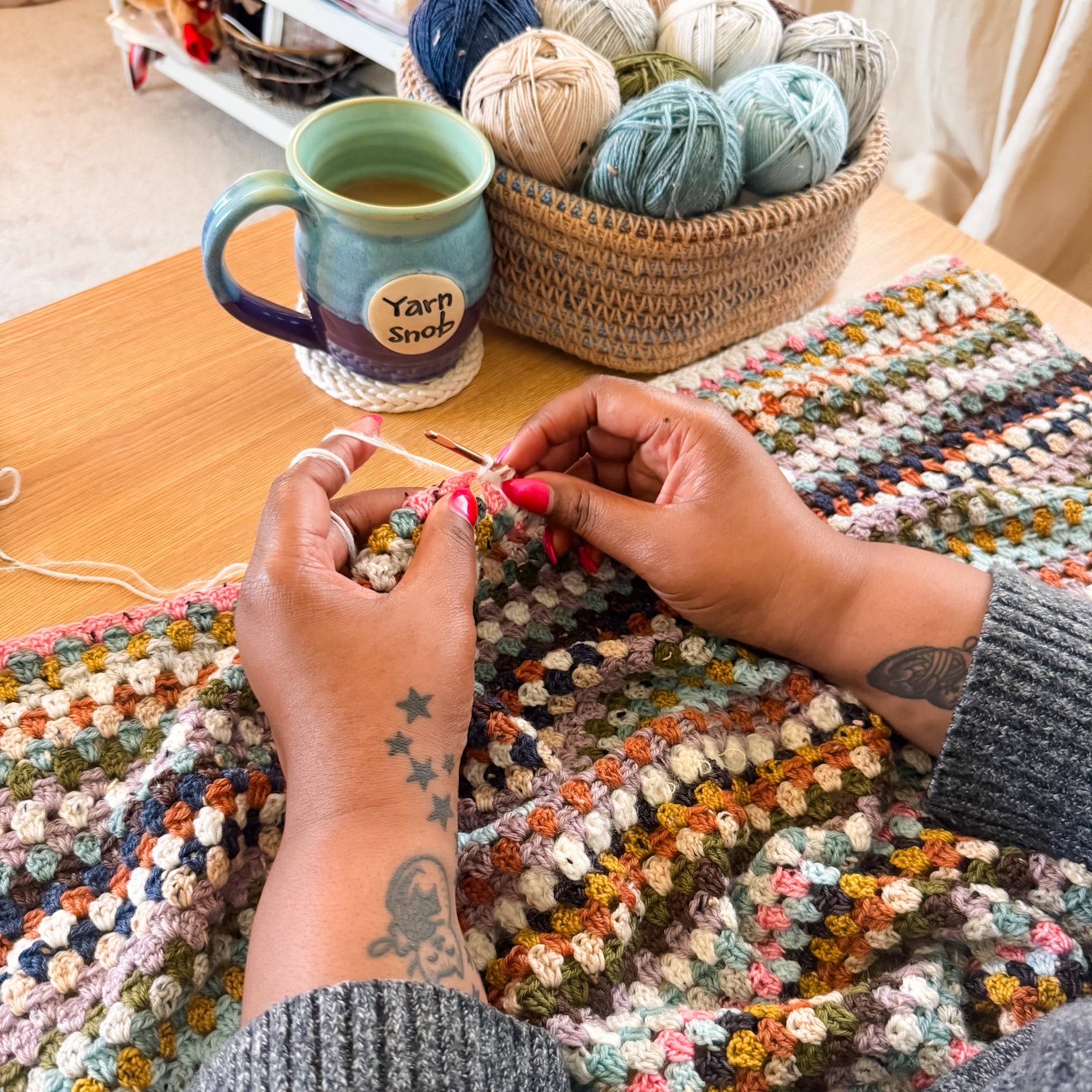 Hands crocheting a colorful blanket beside a yarn-filled basket and a mug.