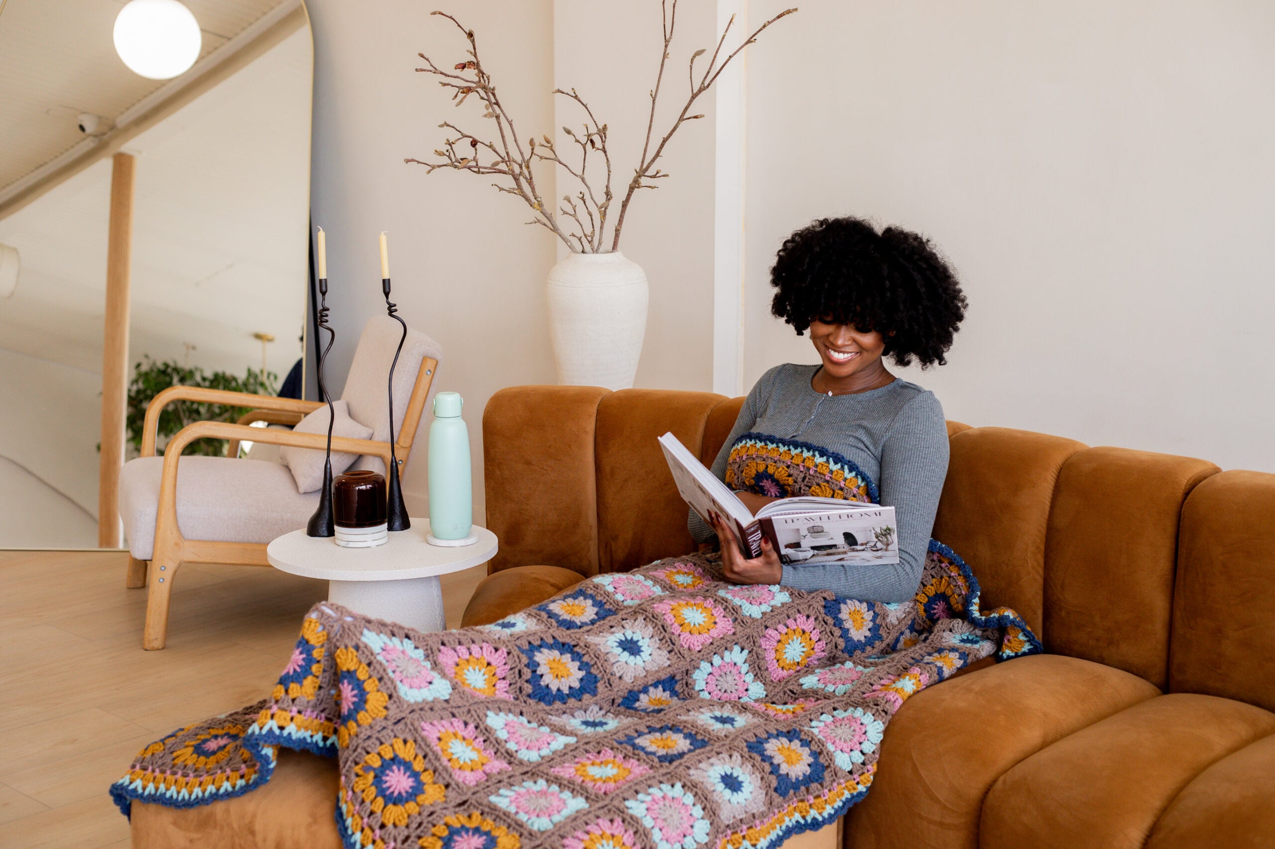 Woman on couch reading a large book tucked under a colorful handmade crochet throw blanket - make the Woodstock Granny Blanket using my free tutorial and pattern