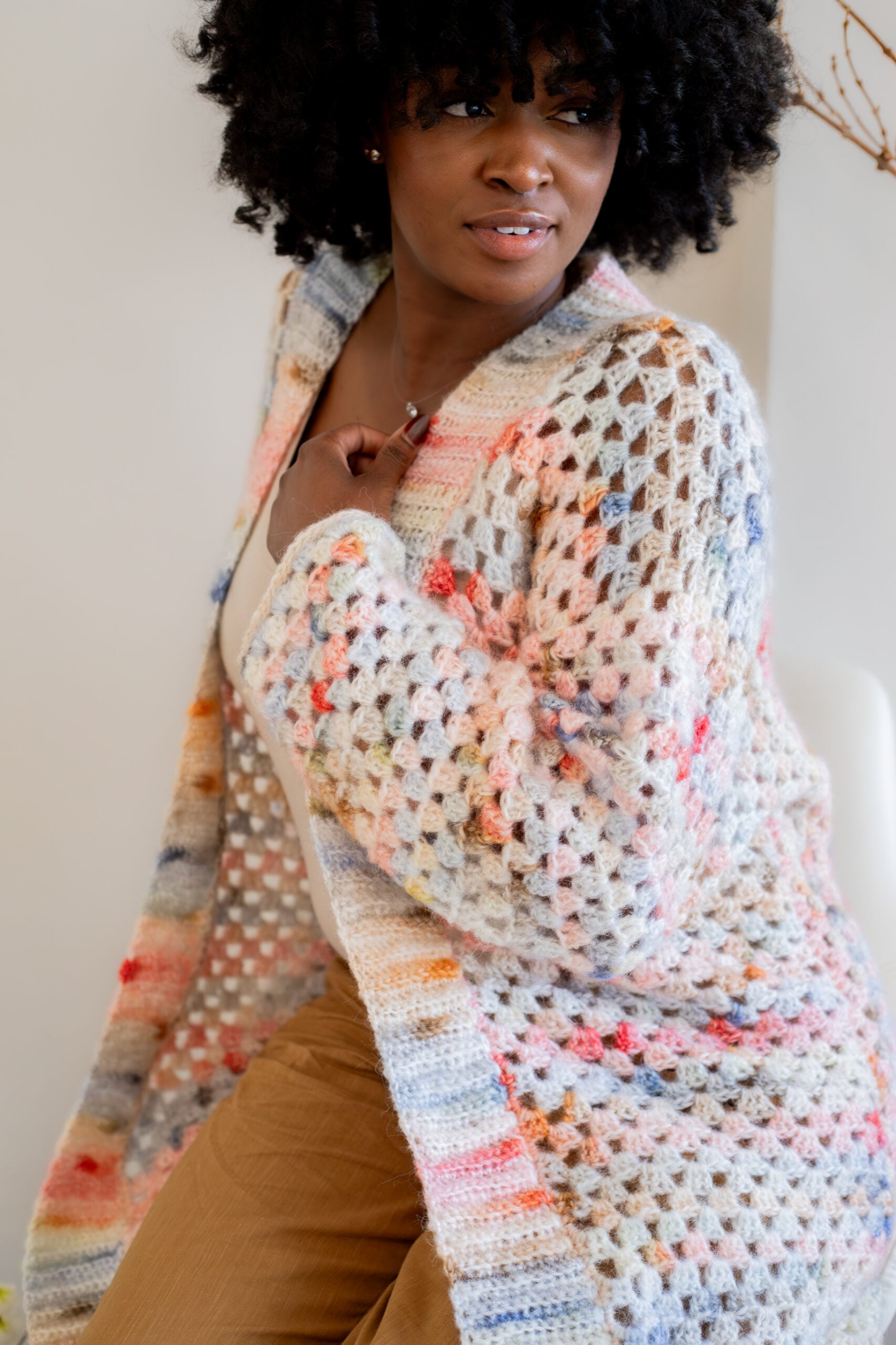 Woman sitting, wearing a pink and blue granny square cardigan - free crochet pattern