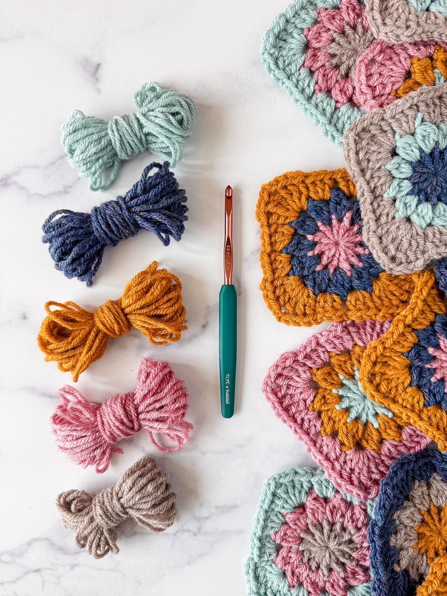 Colorful granny squares sitting next to leftover yarn with tlyarncrafts x hobbii crochet hook in the middle