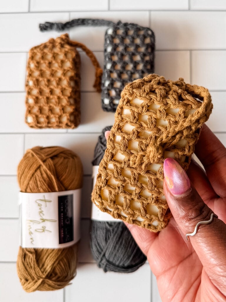 Tan and charcoal Easy Peasy crochet soap savers displayed with wrapped soap bars and Lion Brand Touch of Linen yarn.