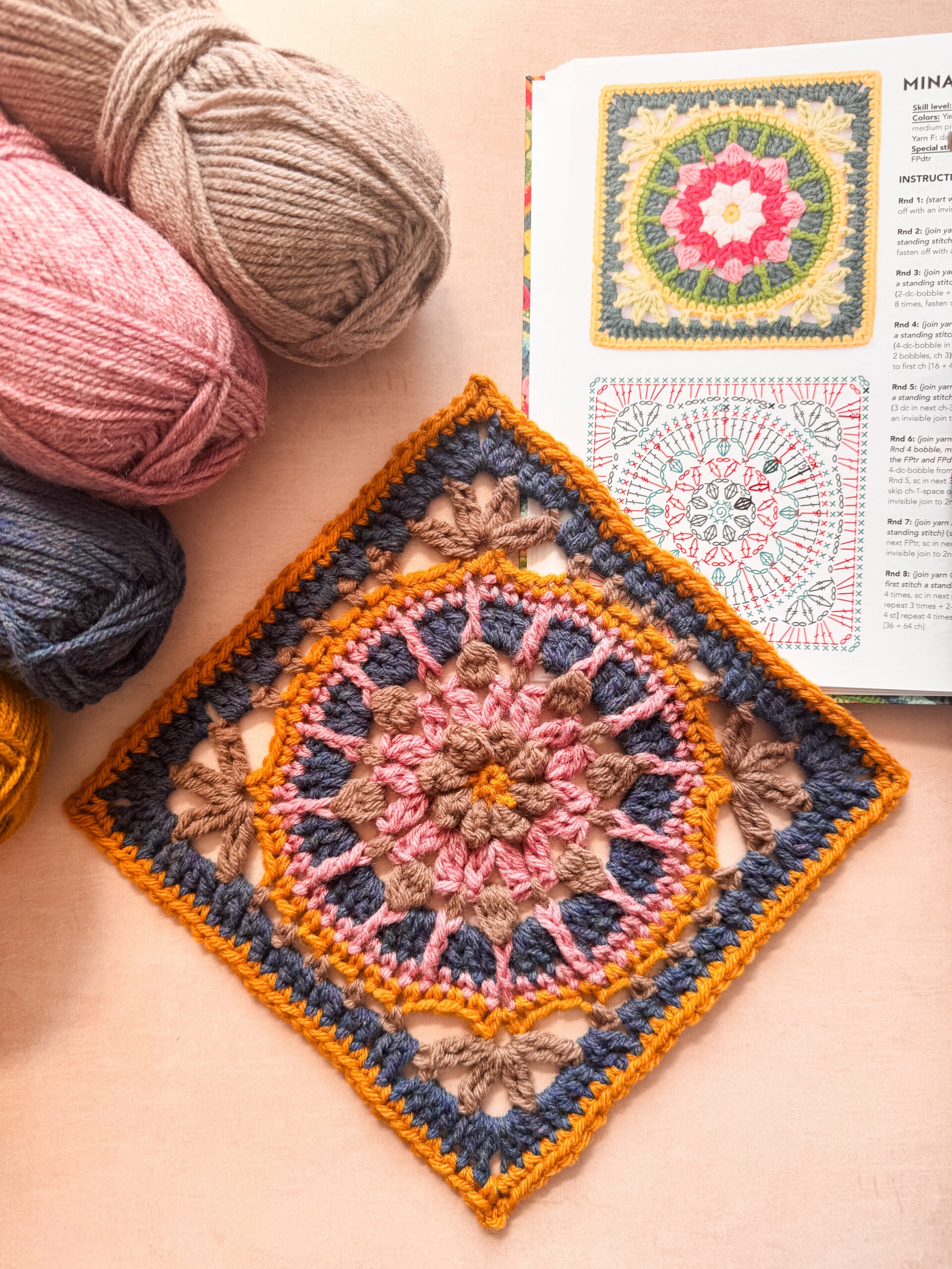 IMG_9814 Colorful crocheted Mina Square beside yarn and an open book showing the finished motif and its crochet symbol chart.